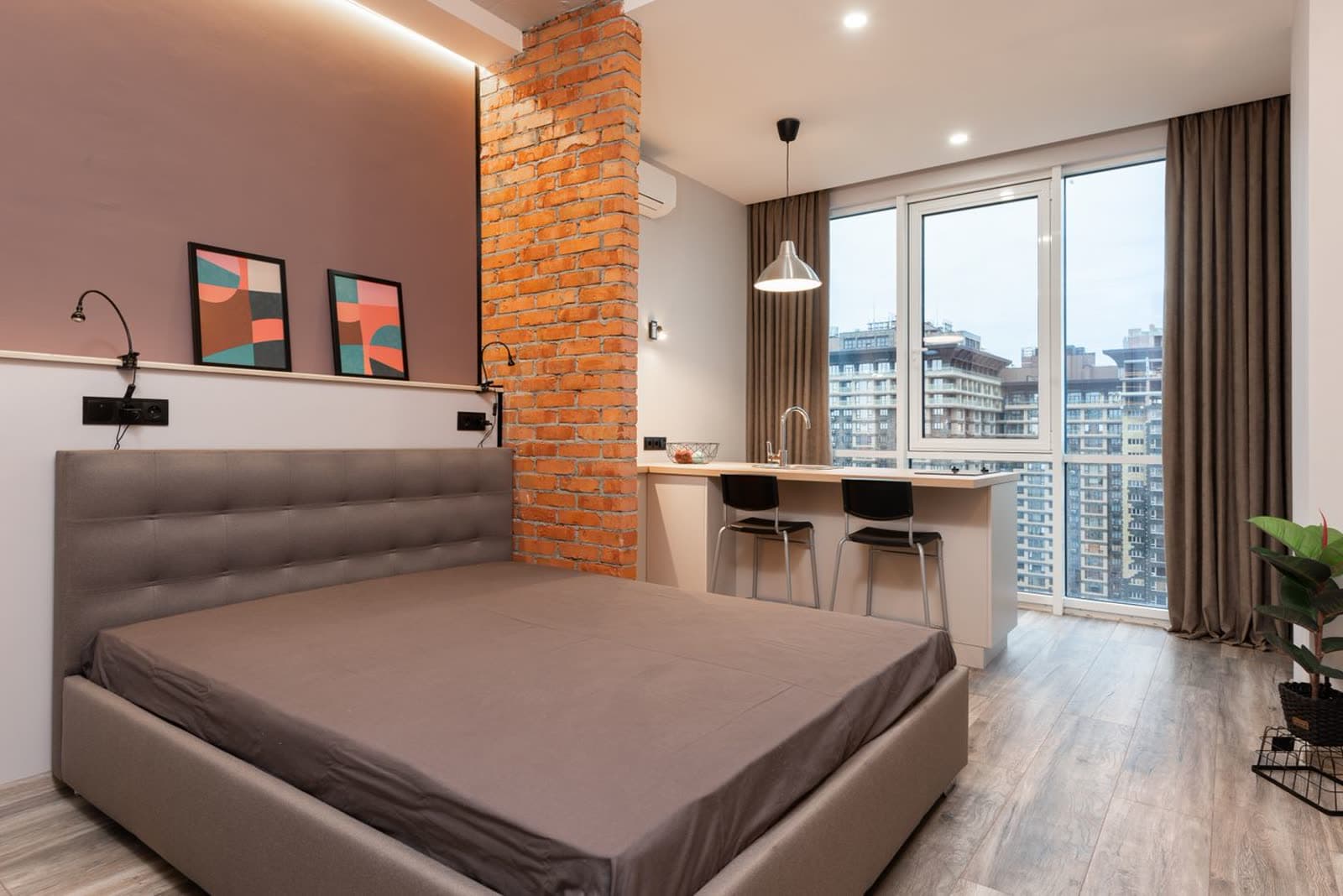 Studio mobiliado com cama de casal, parede de tijolos e bancada, com janela ampla para várias torres residenciais
