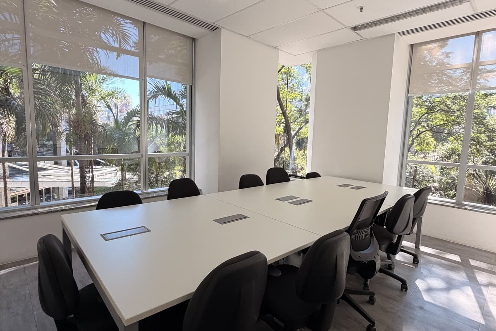 Sala de reunião clara com mesa branca, cadeiras e janelas amplas para a vegetação